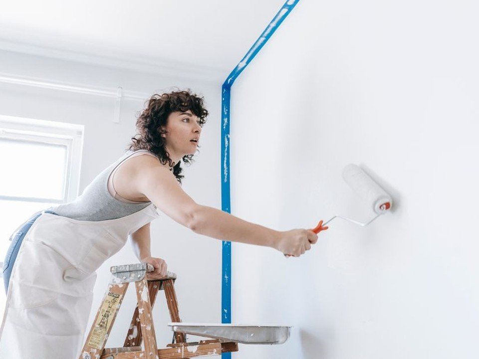 Artisan peintre appliquant au rouleau une teinte choisie sur le grand mur du séjour d'un pavillon des années 1960 habité par son propriétaire à Bron, projet de décoration mené pièce par pièce